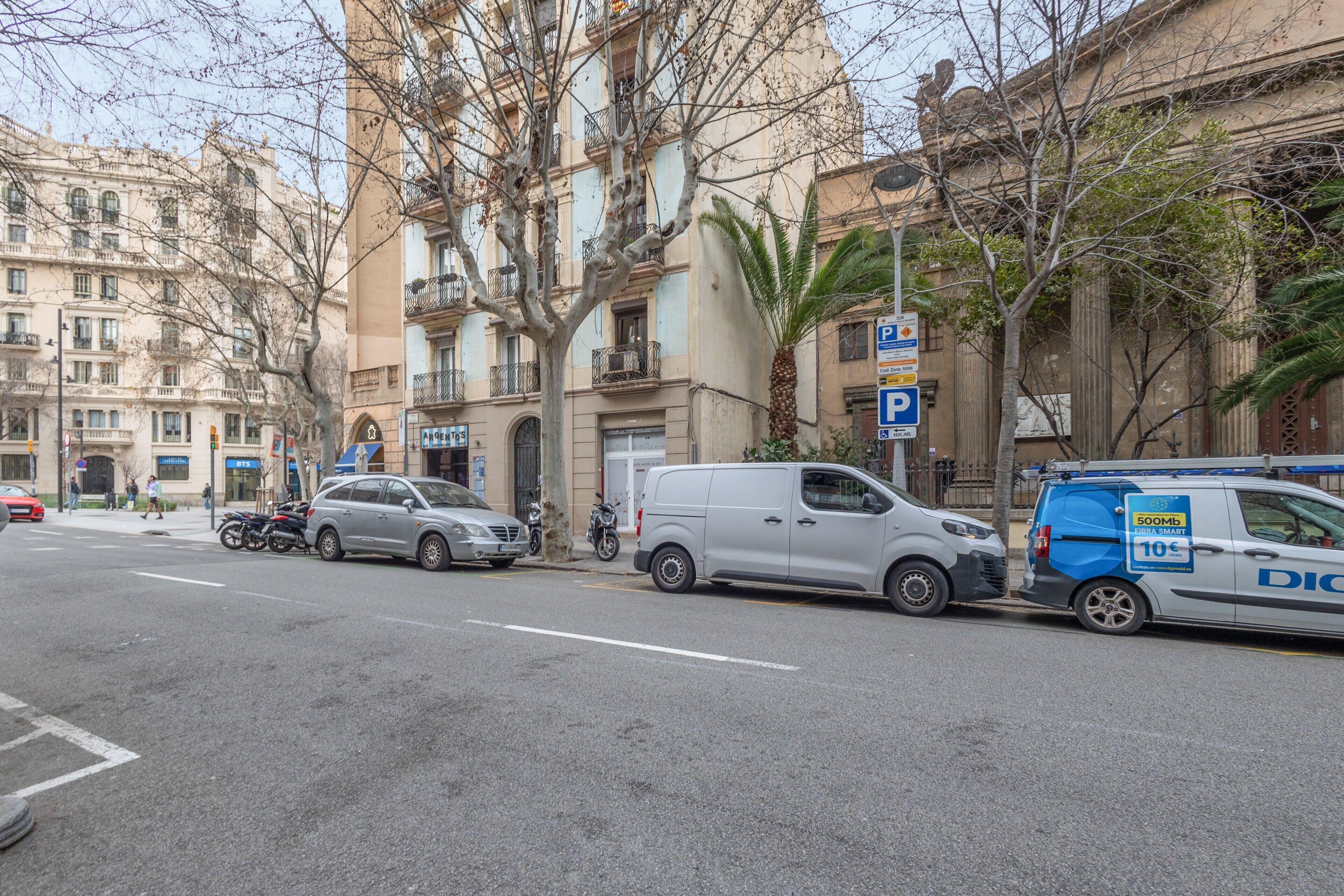 A street with parked cars, a van, and a building with balconies.