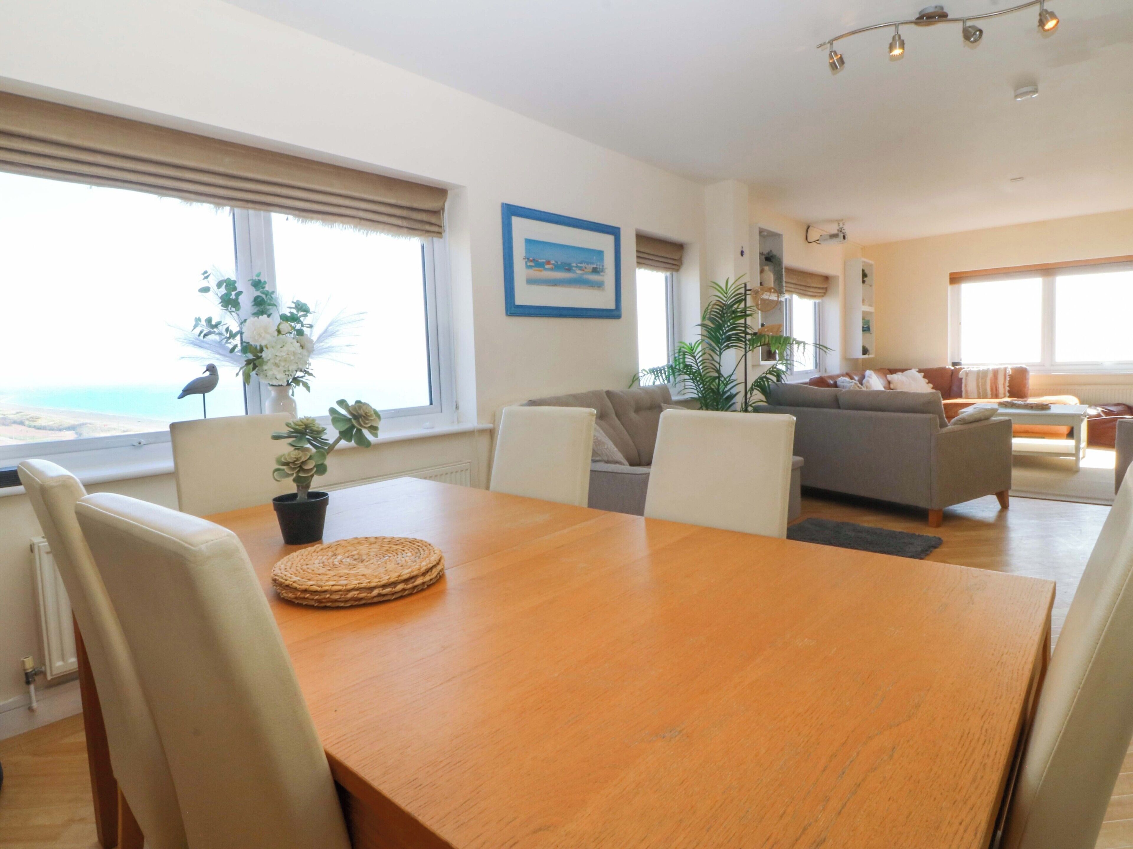 Een moderne woonkamer met een houten eettafel, witte stoelen, een grijze bank en uitzicht op zee door grote ramen.