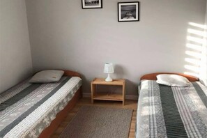 Ein kleines, schlichtes Zimmer mit zwei Einzelbetten, einem Nachttisch mit Lampe sowie zwei gerahmten Bildern an der Wand.