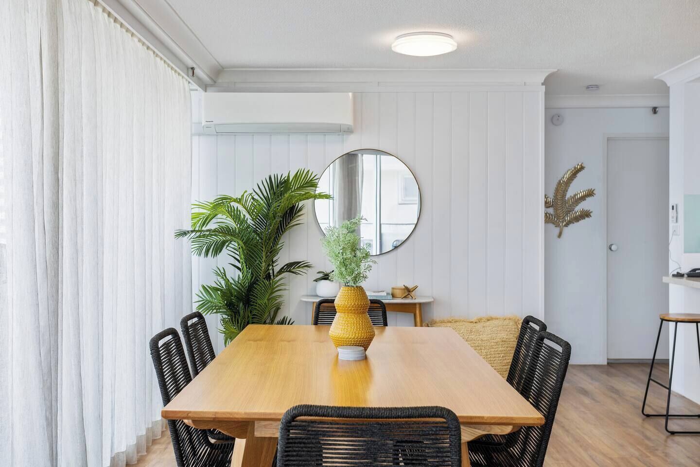 A dining area with a wooden table, black wicker chairs, a large potted plant, and a round mirror on the wall.