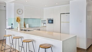 A modern kitchen with white cabinetry, a central island with stools, a clock, and built-in appliances. - Oceans 14th Beachfront Resort style Living (Broadbeach)
