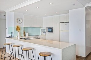 A modern kitchen with white cabinets, a central island with stools, a microwave, and a refrigerator. - Oceans 14th Beachfront Resort style Living (Broadbeach)