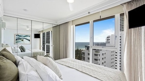 A hotel room with a large bed, a ceiling fan, a TV, and a view of the city and ocean. - Oceans 14th Beachfront Resort style Living (Broadbeach)