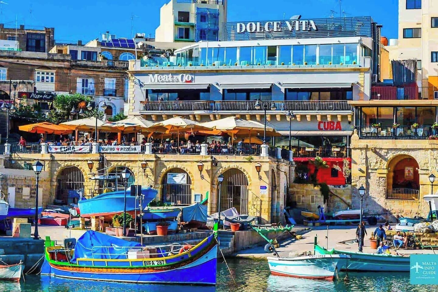 A waterfront building with 'Dolce Vita' and 'Meat & Co' signs, a 'Cuba' restaurant, and boats docked in the water.
