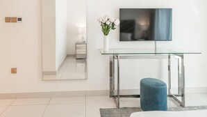 En moderne stue med et glasbord, en blå skammel, et fladskærms-tv monteret på væggen og en vase med hvide blomster.