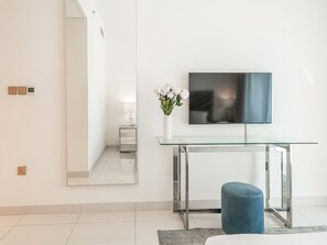 Ein modernes Wohnzimmer mit einem Glastisch, einem blauen Ottomanen, einem an der Wand montierten Flachbildfernseher und einer Vase mit weißen Blumen.