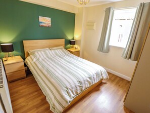 A bedroom with a bed, two bedside tables, a nightstand, a wall-mounted lamp, a window with curtains, and a painting on the wall. - 3 The Old Drill Hall (Alford)