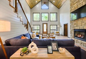 A spacious living room with a large sectional sofa, a fireplace, and a staircase leading to an upper level. - Hot Tub | Fire Pit | Foosball | Arcade Machine (Broken Bow)