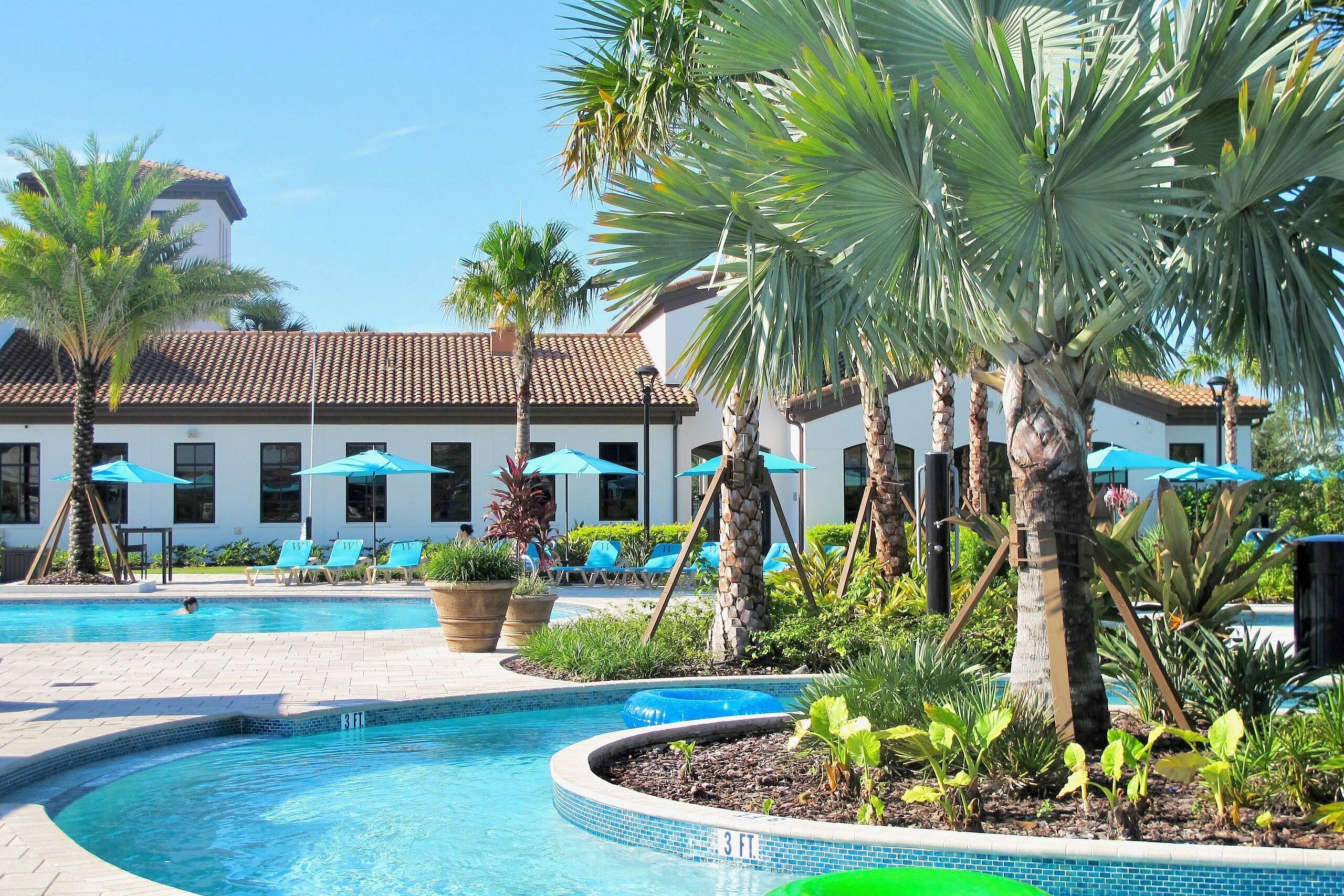 A swimming pool with lounge chairs and umbrellas, surrounded by palm trees and a building with a tiled roof.