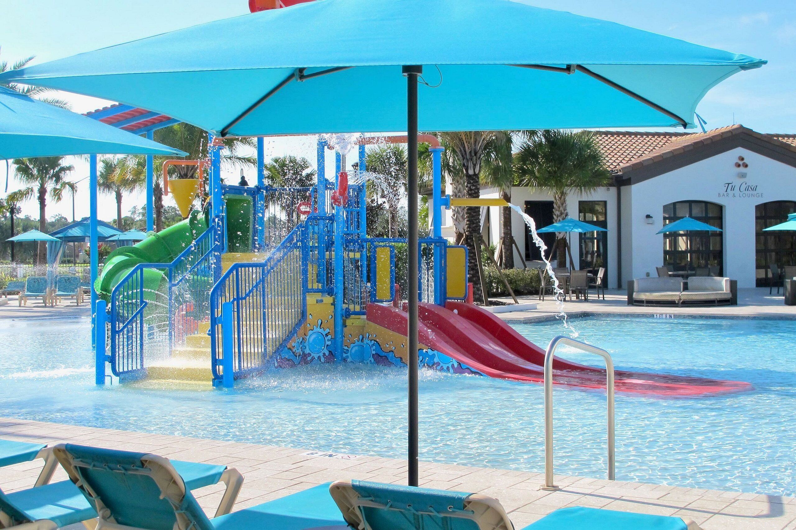 A pool area with a water play structure, lounge chairs, and a bar/lounge building.
