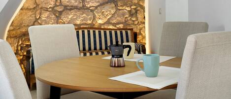 A round wooden table with a stone wall backdrop, a coffee maker, and two mugs.