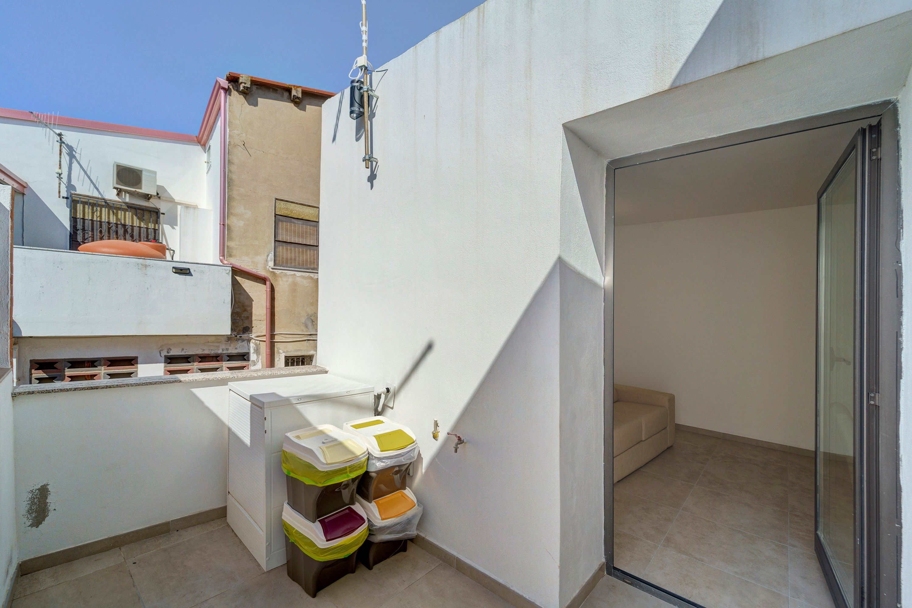 A small, enclosed balcony area with a white wall, a metal railing, and a door leading to an adjacent room. There are two trash bins with colorful lids.