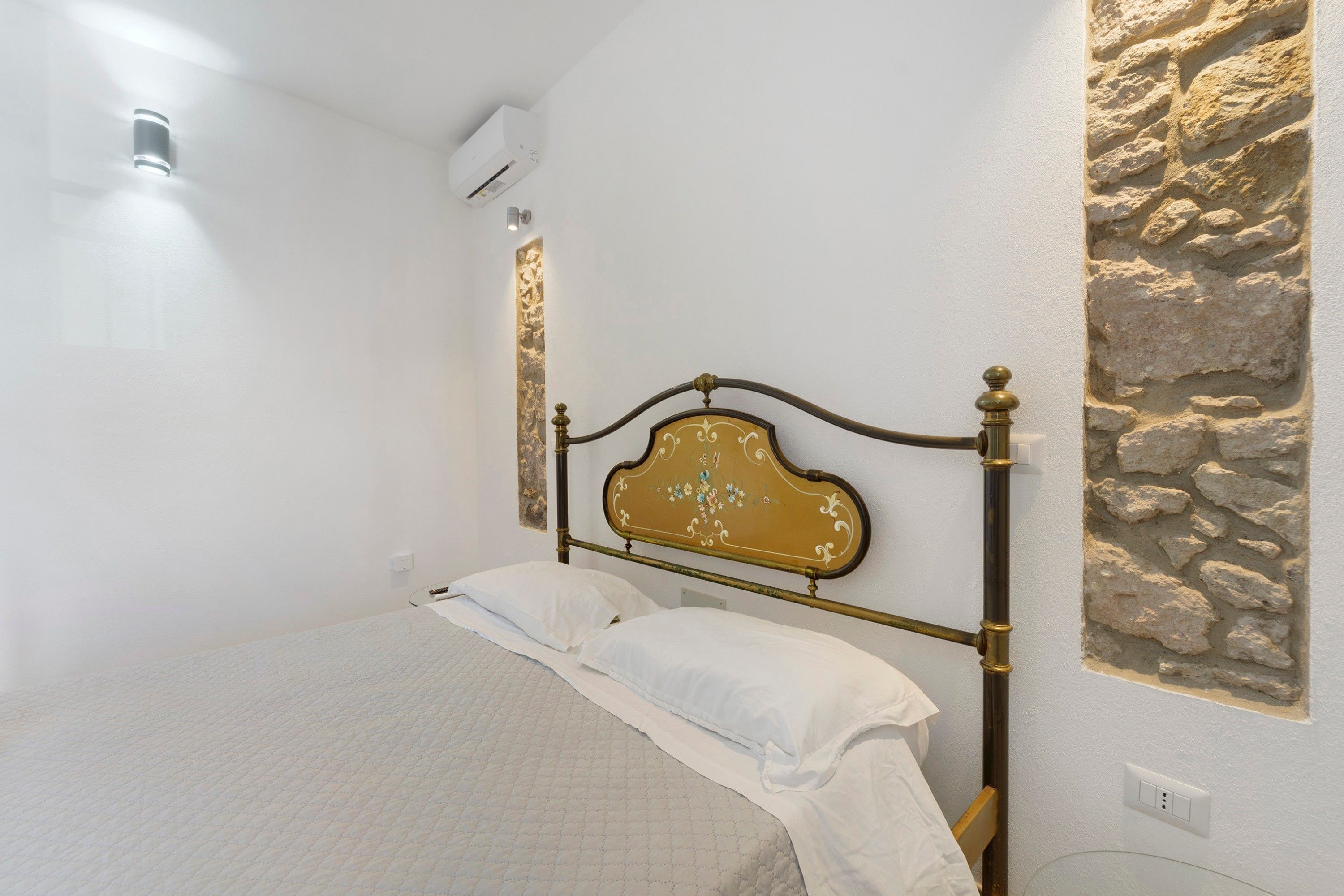 A bed with a headboard, a wall-mounted air conditioner, and a stone wall.