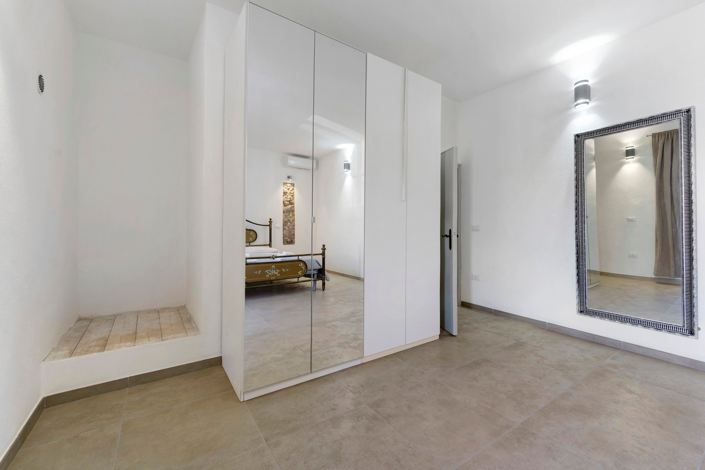A room with a bed, a wardrobe, a mirror, and a tiled floor.