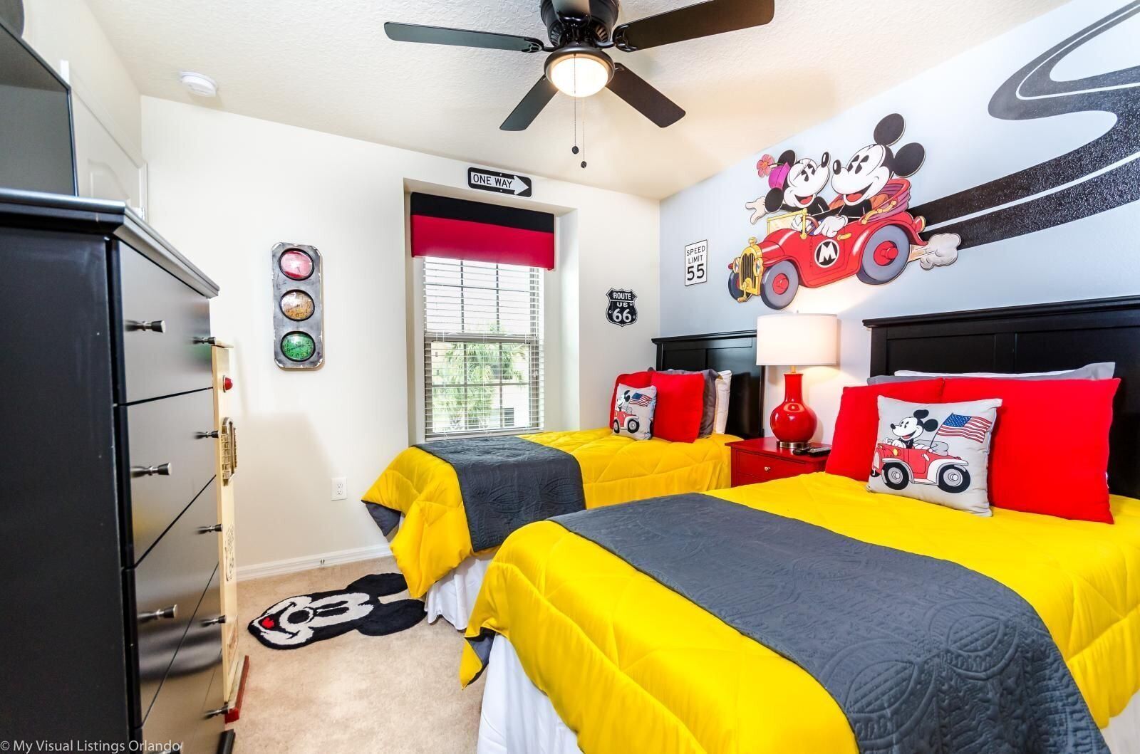 A hotel room with two beds, a ceiling fan, a red and white wall mural, and a window with blinds.