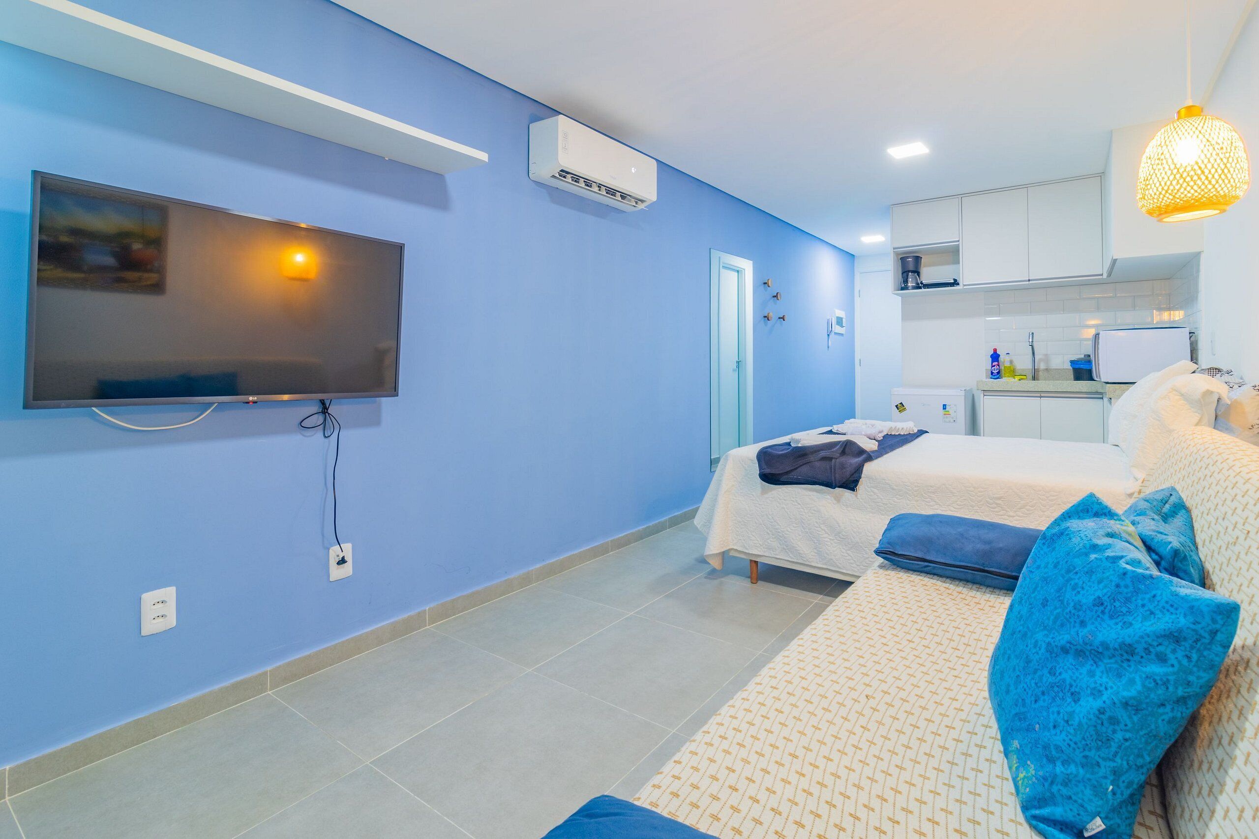 A bedroom with a TV, air conditioning unit, and a kitchenette in the background.