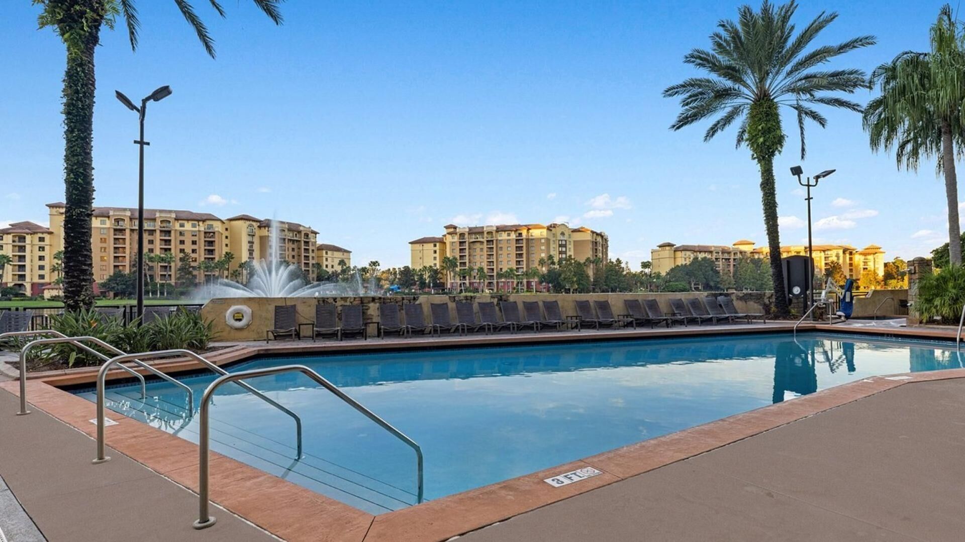 A swimming pool with a water fountain, surrounded by palm trees and lounge chairs, with hotel buildings in the background.