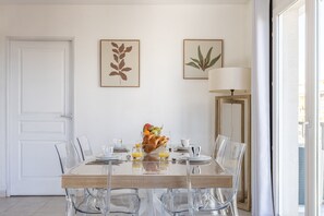 A dining area with a wooden table, clear chairs, and a fruit bowl. There are framed botanical prints on the wall and a lamp on the right. - Duplex 2 Chambres -balcon-parking- Carnota2b269 (Cannes)