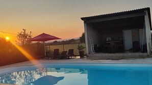 Una piscina con sillas de descanso, un área techada con una escalera, y una puesta de sol al fondo.