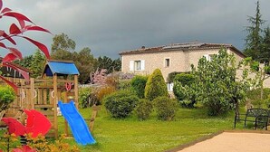 Un parque infantil con un tobogán azul y una casita de madera, rodeado de césped y un edificio con tejado de tejas.