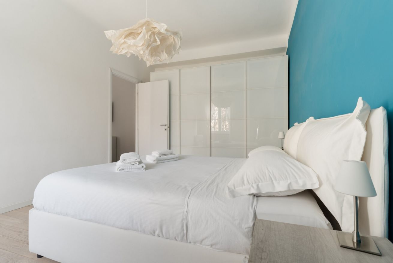 Une chambre moderne avec un grand lit, du linge de lit blanc, une lampe de chevet et un mur d’accent bleu.