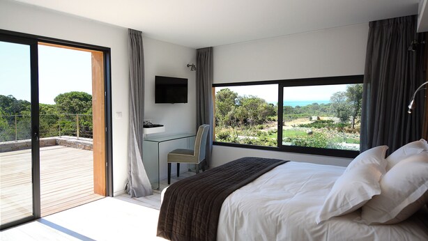 A bedroom with a large bed, a desk, and a TV. It has a balcony with a view of greenery and a building. - Architect-designed 4-bedroom villa with pool in palombaggia with sea view (Porto-Vecchio)
