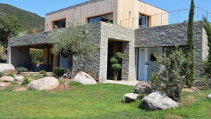 A modern house with a stone facade, wooden upper levels, and a well-maintained garden with large rocks and shrubs. - Architect-designed 4-bedroom villa with pool in palombaggia with sea view (Porto-Vecchio)
