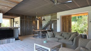 A spacious living area with a wooden floor, a sofa, a coffee table, and a television. There is a staircase leading to another level, and a large window providing a view of greenery outside. - Architect-designed 4-bedroom villa with pool in palombaggia with sea view (Porto-Vecchio)