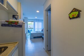 En smal hall med ett köksutrymme på vänster sida, en säng med ett blått överkast i mitten och en dörr som leder till ett sovrum på höger sida.