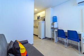 Ein kompakter Wohnraum mit Küchenzeile, Wasserspender, Mikrowelle und Kühlschrank. Zwei blaue Stühle und ein graues Sofa mit einem bunten Kissen.