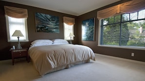 A bedroom with a large bed, two bedside tables, and two lamps. There are two large windows with curtains, and two paintings on the wall. - King bed, en-suite bathroom w/separate tub/shower (Vancouver)