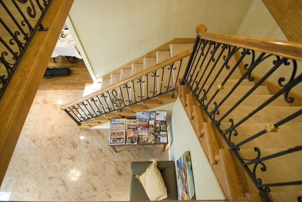 Un escalier avec une rampe en bois et des balustres en métal noir. En contrebas, on aperçoit une table sur laquelle sont posées des brochures et une guitare.
