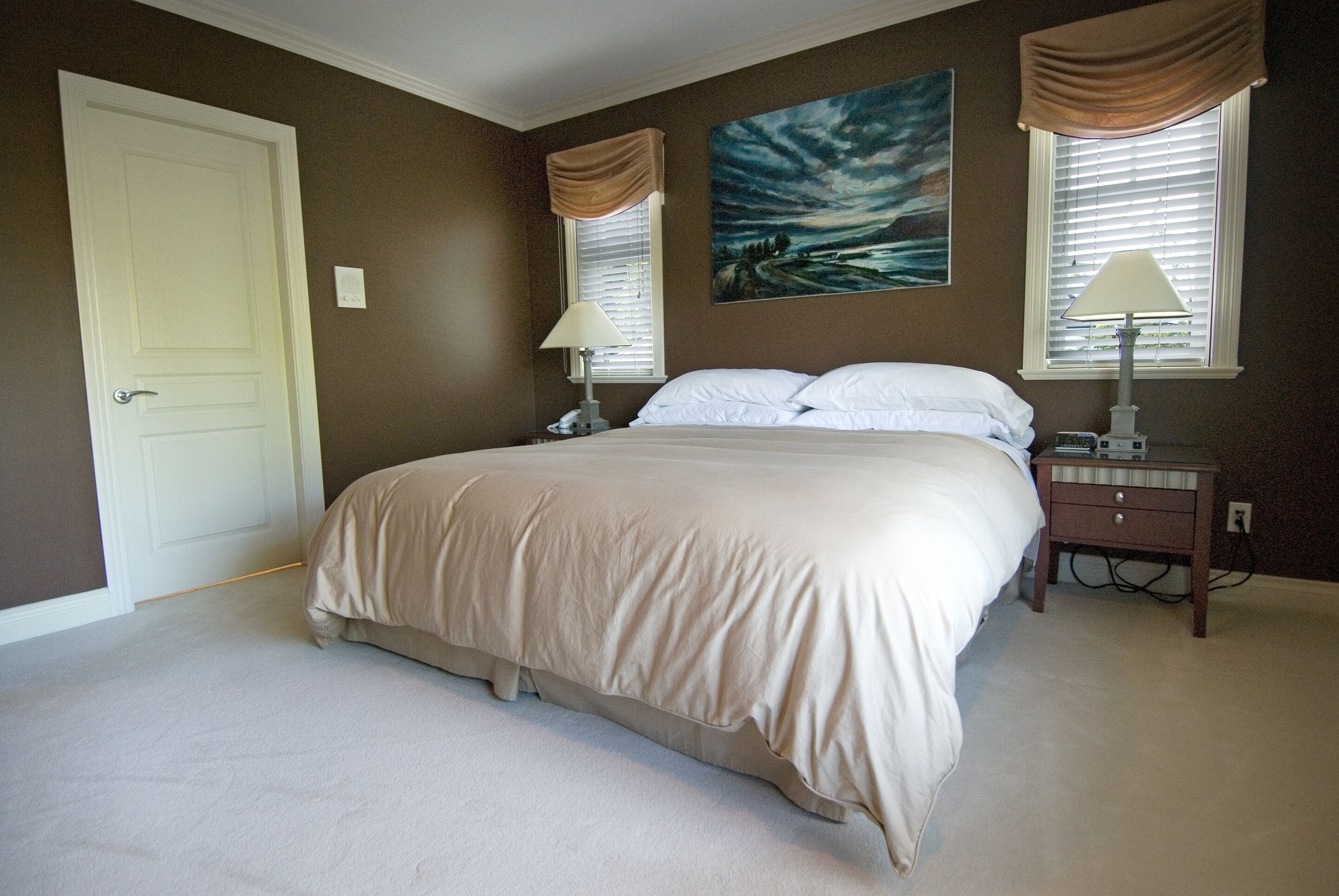 A bedroom with a large bed, two bedside tables, a nightstand, a door, and a painting on the wall.
