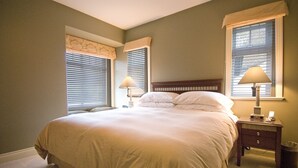 Ein Schlafzimmer mit einem großen Bett, zwei Nachttischlampen, einem Nachttisch und einem Fenster mit Jalousien.