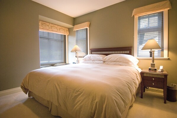 A bedroom with a large bed, two bedside lamps, a nightstand, and a window with blinds.