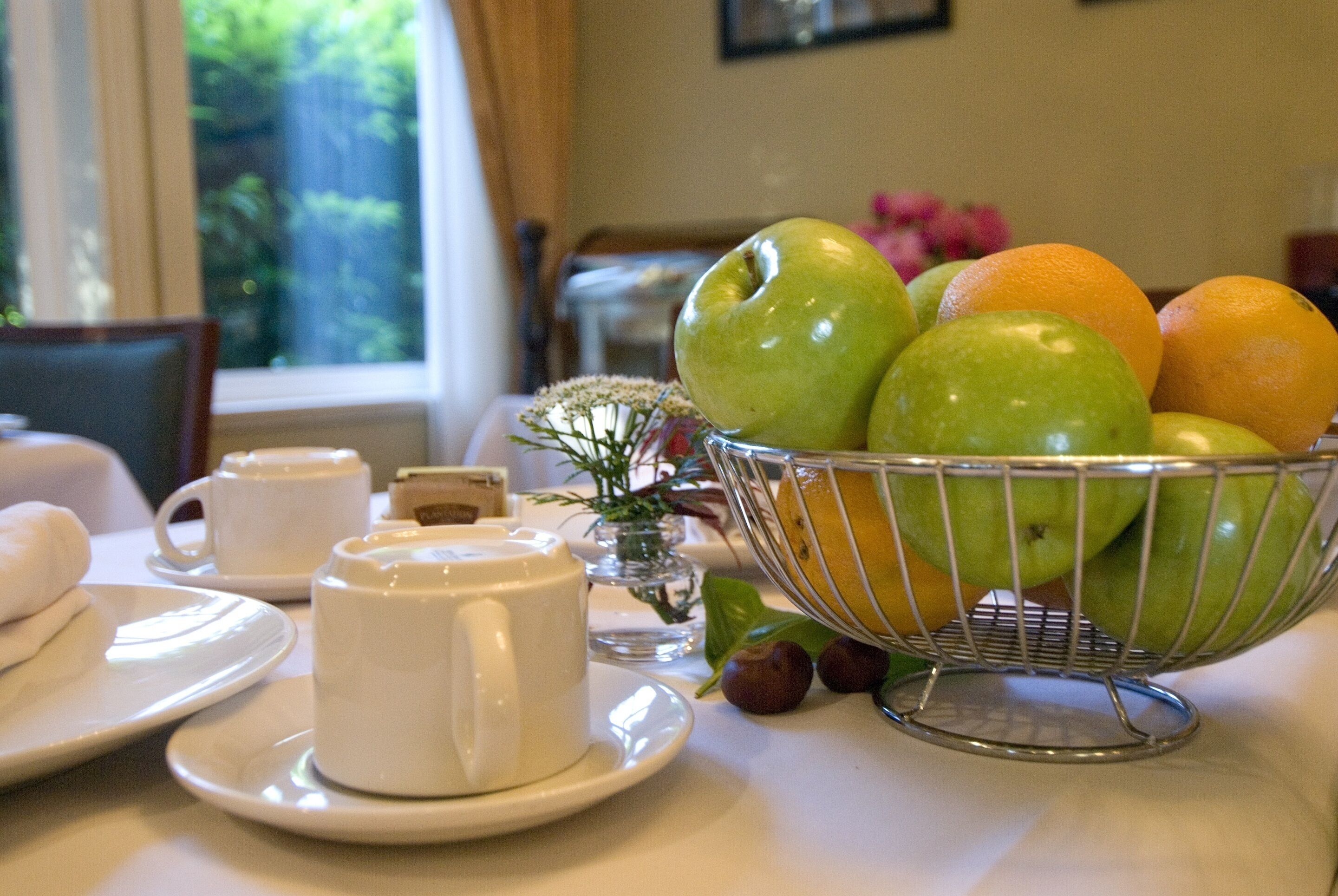 Una mesa de comedor con un frutero que contiene manzanas y naranjas, una taza de café blanca y un jarrón pequeño con flores.