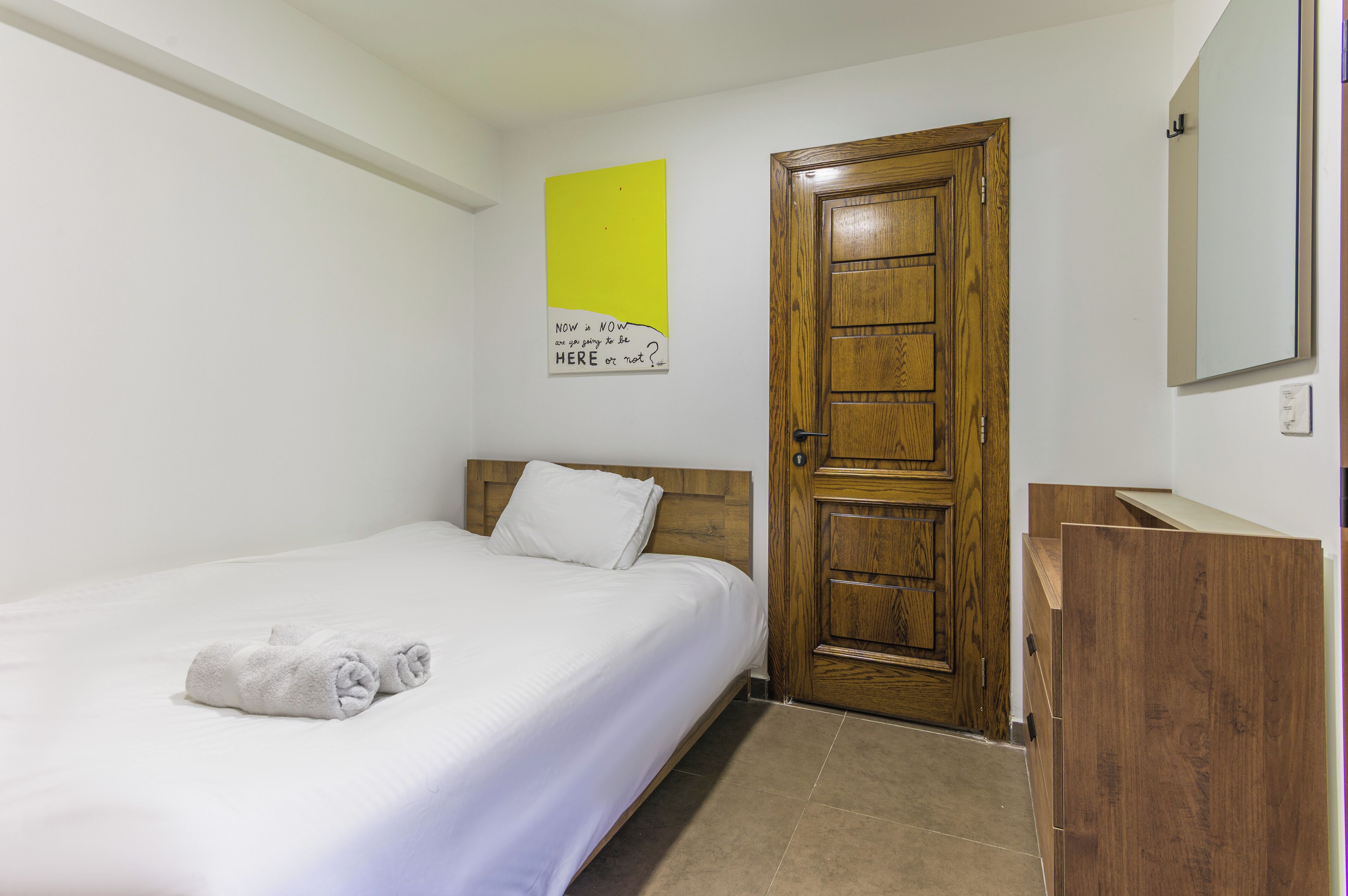 A hotel room with a single bed, a wooden door, a small wooden cabinet, and a wall-mounted mirror.