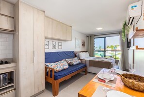A compact living space with a kitchenette, a sofa with floral cushions, a dining table with wine glasses, and a view of the outdoors. - Villadelmar 204 - Studio With Balcony by Carpediem (Ipojuca)