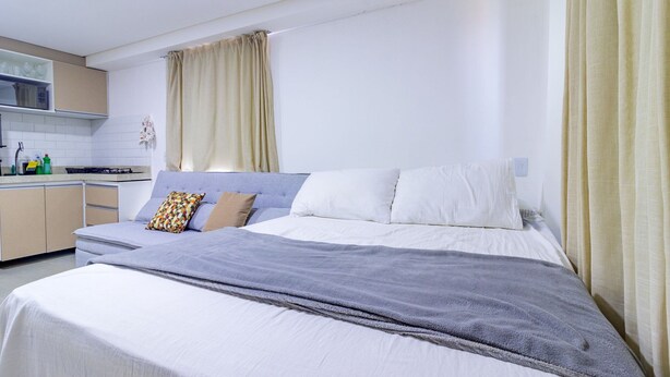 A bedroom with a bed, a bedside table, a window with curtains, and a kitchenette in the background. - Villa Del Mar #203- Flat With Balcony by Carpediem (Ipojuca)