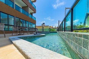 A swimming pool with lounge chairs and a view of a modern building with large windows. - Villa Del Mar #203- Flat With Balcony by Carpediem (Ipojuca)