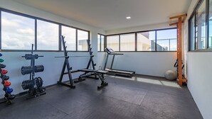 A gym with weightlifting equipment, treadmills, and a large window offering a view of the sky. - Porto Prime #308 - Studio Confort by Carpediem (Ipojuca)