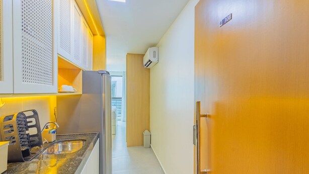 A narrow hallway with a kitchen area on the left, featuring a sink, cabinets, and a refrigerator. A wooden door with a number plate is on the right. - Porto Prime #308 - Studio Confort by Carpediem (Ipojuca)