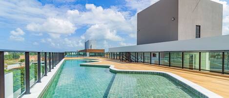 A modern swimming pool with a wooden deck, surrounded by glass railings and a clear sky.