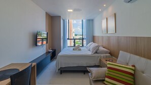 A modern hotel room with a large bed, a TV, a desk, and a view of buildings through the window. - Porto City #314 - Porto de Galinhas by Carpediem (Ipojuca)