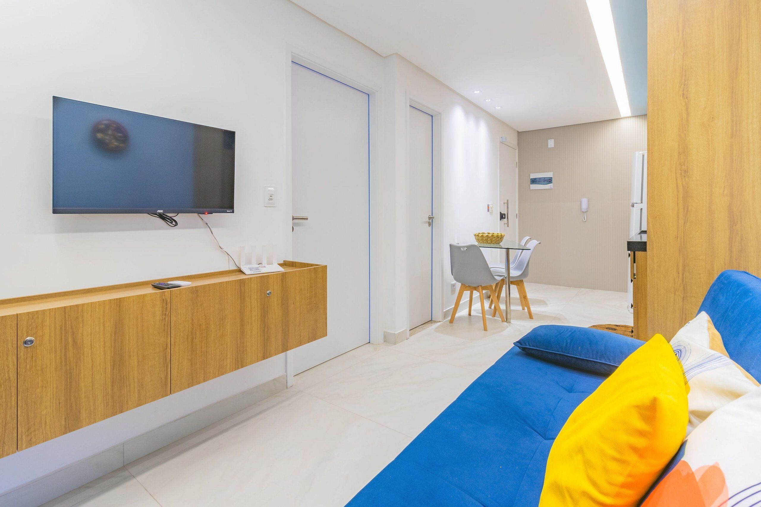 Una habitación moderna con una televisión de pantalla plana montada en la pared, una mesita consola de madera, un sofá azul con cojines de colores, una pequeña mesa de comedor con sillas y una puerta de madera.