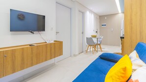 Ein modernes Zimmer mit einem wandmontierten Flachbildfernseher, einem Holzkonsolentisch, einem blauen Sofa mit bunten Kissen, einem kleinen Esstisch mit Stühlen und einer Holztür.