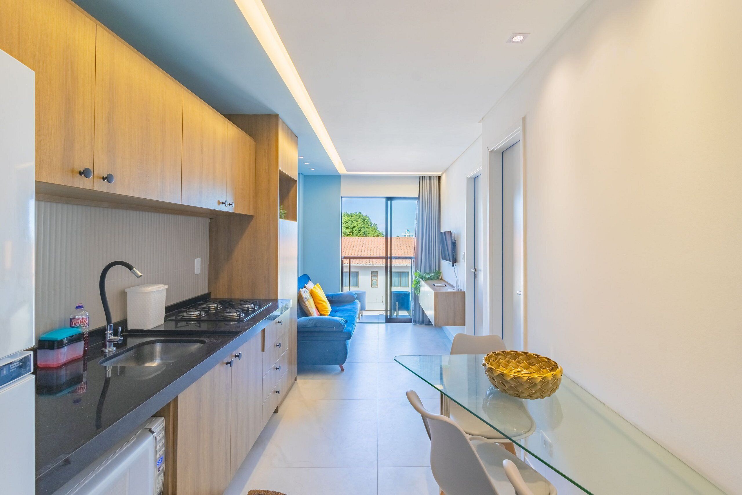 Una cocina moderna con encimera negra, estufa y fregadero. En la zona de estar, hay un sofá azul y una mesa de vidrio con sillas.