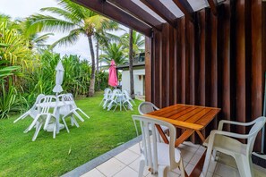 A patio area with a wooden table and white plastic chairs, surrounded by lush greenery and palm trees. - Beach Class #7113- Bungalow by Carpediem (Ipojuca)