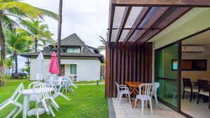 A patio with white plastic chairs and a wooden table, a covered area with a glass wall, and a building with a tiled roof and a red umbrella. - Beach Class #7113- Bungalow by Carpediem (Ipojuca)