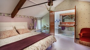 Un dormitorio con una cama, una silla roja, una mesita de noche, una lámpara y un baño visible a través de una puerta abierta.